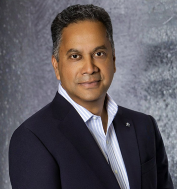 Professional head-and-shoulders portrait of a man in a dark blazer and light striped shirt against a textured gray backdrop.