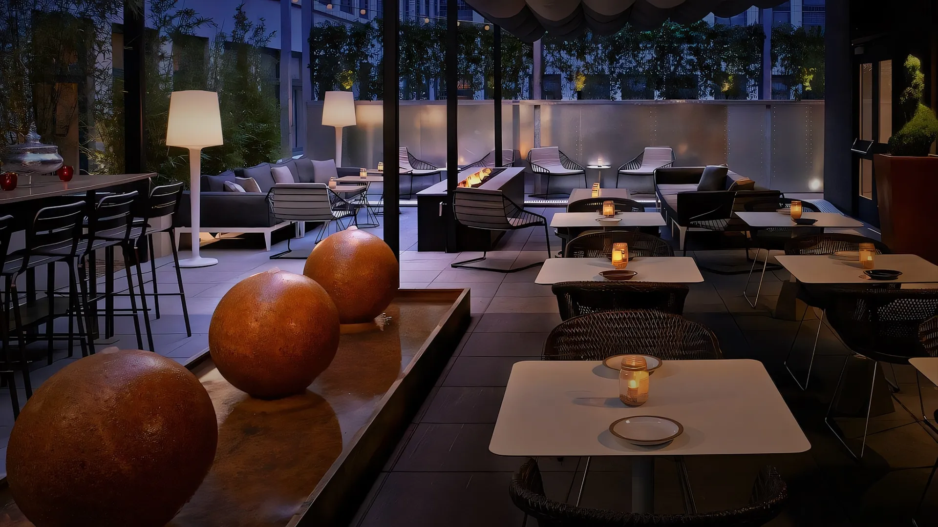 Nighttime outdoor dining terrace with white tables, candles, and modern seating along a water feature with orange spheres.